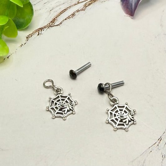 Silver spiderweb dangle earrings with black post backings, displayed on a light marble surface with decorative green leaves.