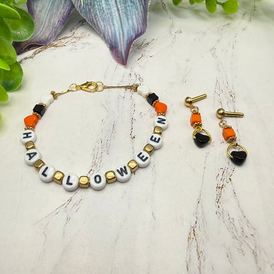 A Halloween-themed beaded necklace and matching drop earrings displayed on a light marble surface with decorative green leaves in the background. The necklace features white letter beads spelling 'HALLOWEEN', accented with black, orange, and gold beads.