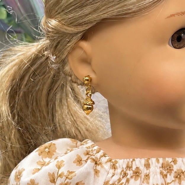 A close-up of a doll wearing a small gold dangling earring with a bead and acorn motif, against a softly blurred background.