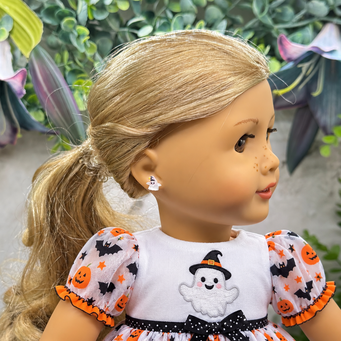 A blonde doll with a ponytail wearing a white Halloween dress with orange and black pumpkin, bat, and ghost patterns, along with a small white ghost stud earring.