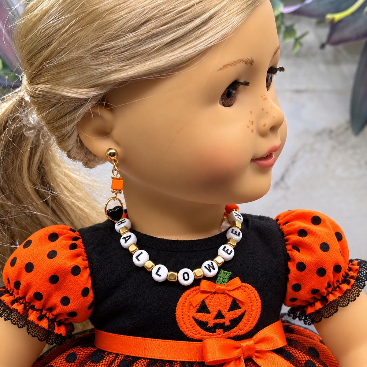 A doll wearing a Halloween outfit with a black top, orange polka-dot sleeves, and an orange pumpkin patch, styled with an orange and white beaded necklace and matching drop earrings.