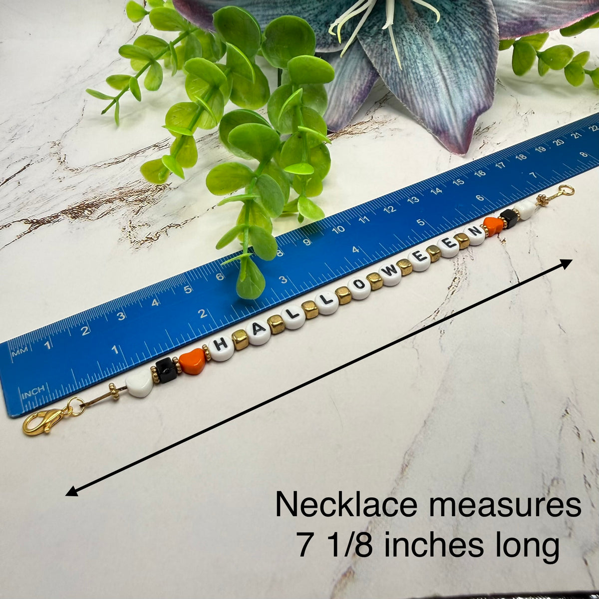 A beaded Halloween-themed necklace spelling 'HALLOWEEN' is shown on a white marble surface with a blue ruler for scale and text reading 'Necklace measures 7 1/8 inches long.' Green leaves and a purple flower are placed around it as styling props.