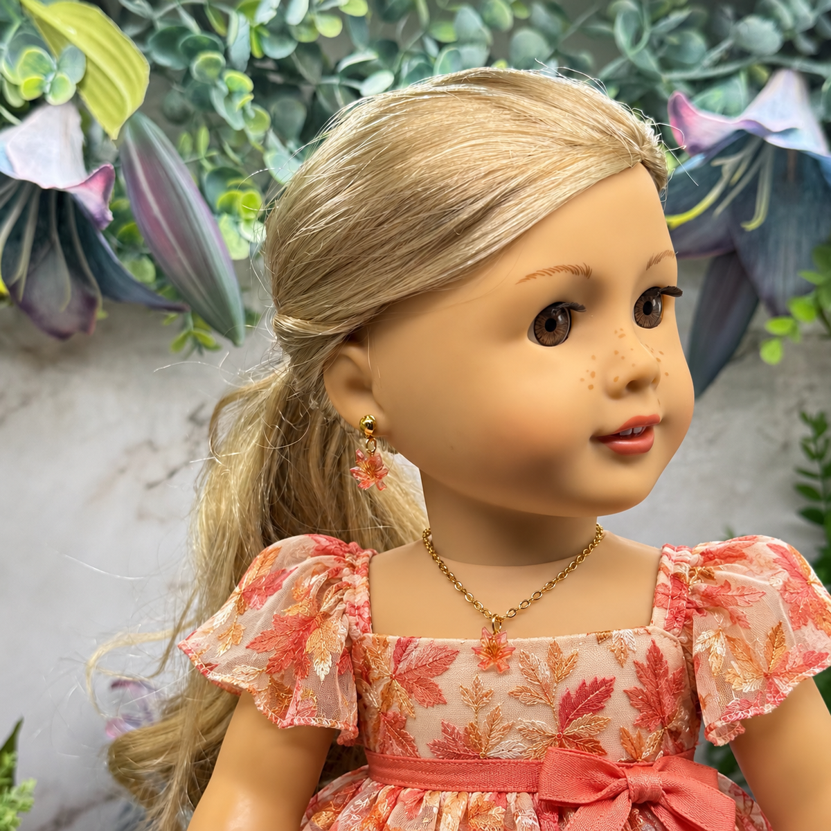 A doll with blonde hair wearing a floral pink and orange dress, gold necklace, and matching earrings, shown against a decorative garden backdrop.