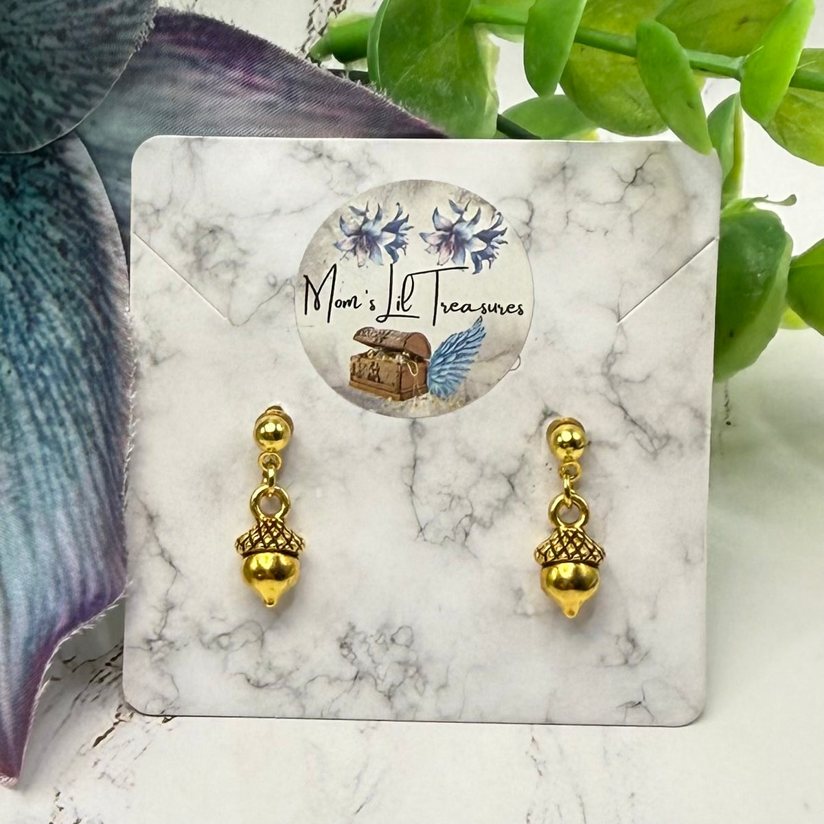A pair of gold acorn drop earrings displayed on a white marble card