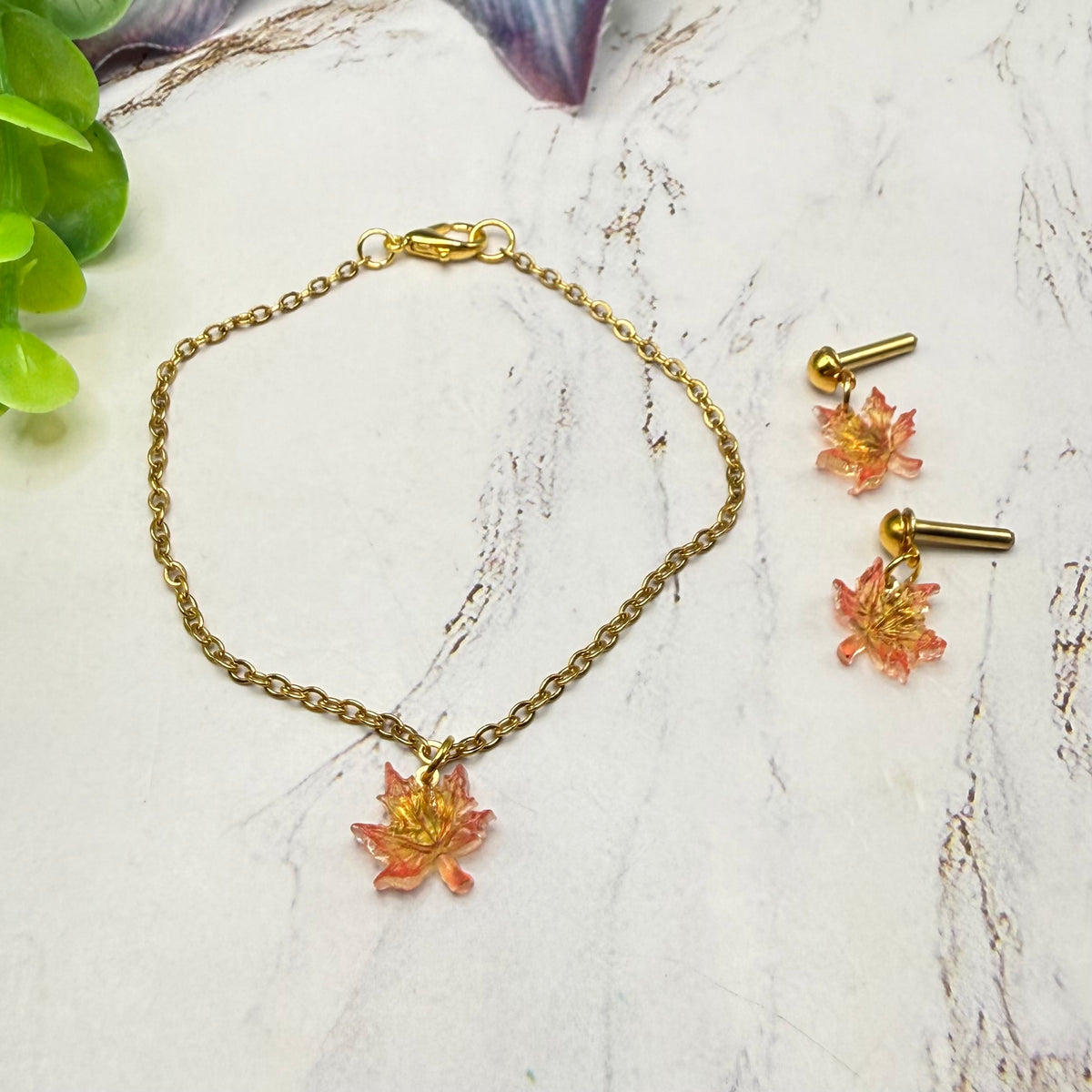 Gold chain necklace and matching stud earrings with small orange-pink maple leaf charms, arranged on a light marble-like surface with green leaves at the corner.