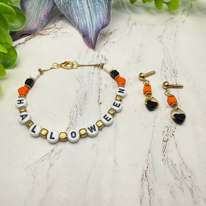 A Halloween-themed beaded necklace and matching drop earrings displayed on a light marble surface with decorative green leaves in the background. The necklace features white letter beads spelling 'HALLOWEEN', accented with black, orange, and gold beads.