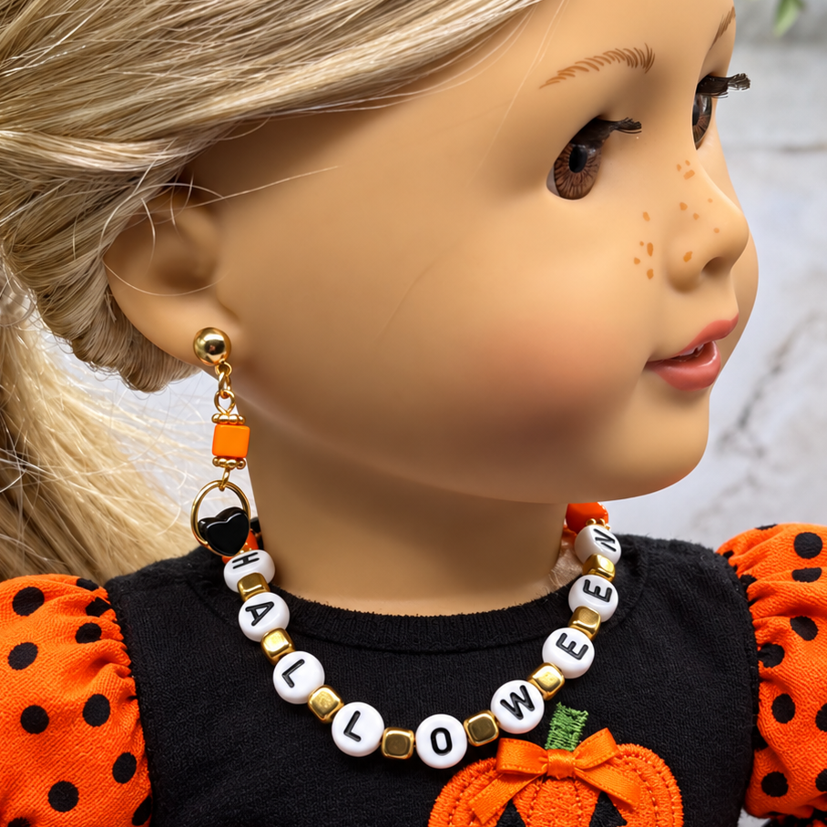 A doll wearing a Halloween-themed beaded necklace and matching drop earring. The necklace has white letter beads spelling HALLOWEEN with gold spacer beads, and the earring is white and orange with a gold heart and ring.