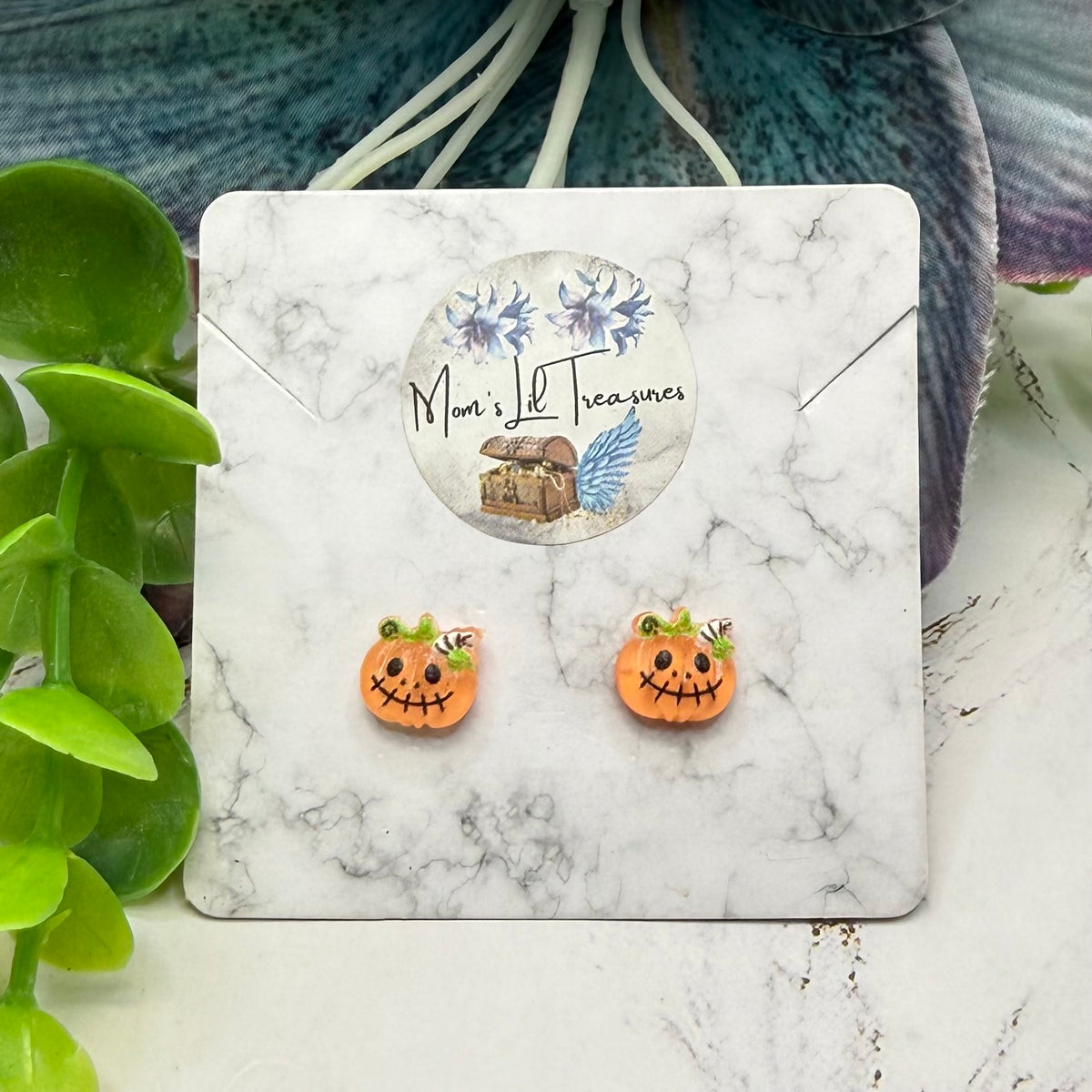 A pair of orange pumpkin-shaped stud earrings with carved jack-o'-lantern faces and small green leaf accents, displayed on a white marble card labeled '