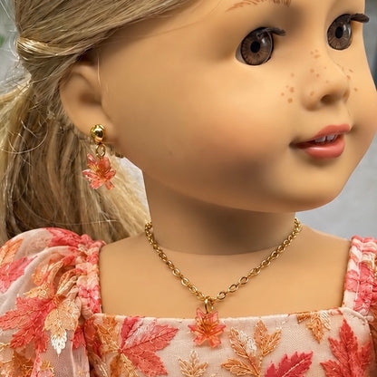 A close-up of a doll wearing a gold leaf drop earring and matching gold leaf pendant necklace, with a pink and orange autumn leaf patterned dress.