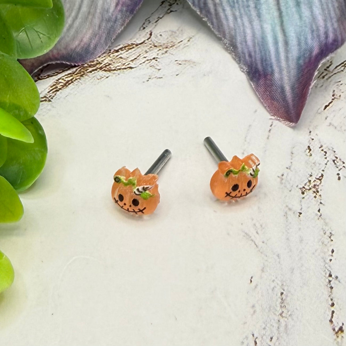 A pair of small orange pumpkin-shaped stud earrings with a cute face and green leaf accents, displayed on a textured white surface with decorative green leaves and a purple fabric in the background.