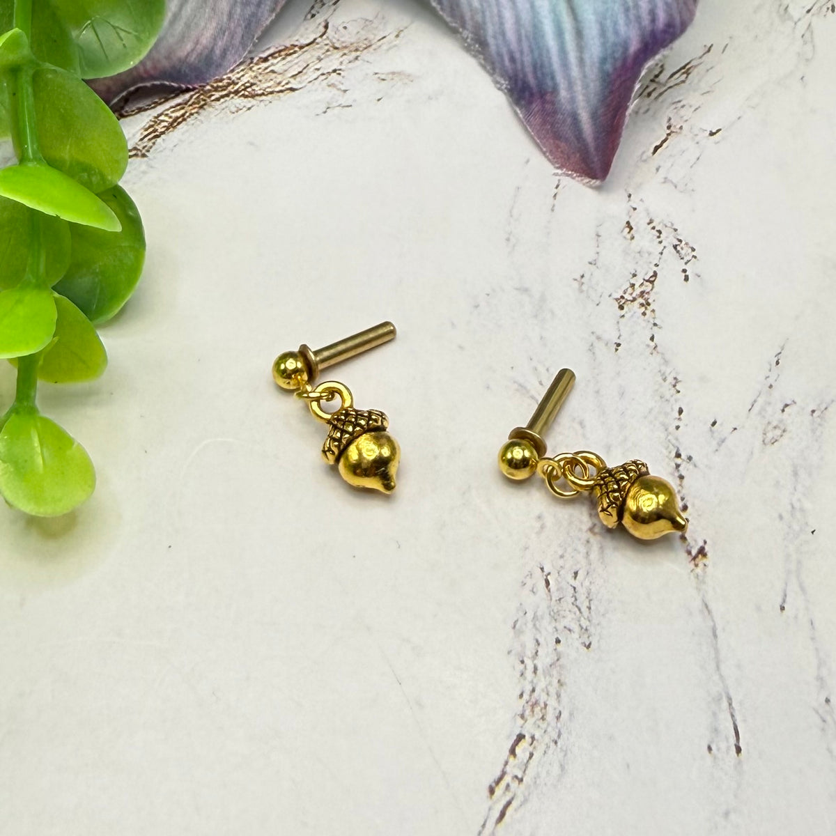 A pair of gold earrings with small dangling acorn charms, displayed on a light textured surface with green leaves and a purple flower petal as props.